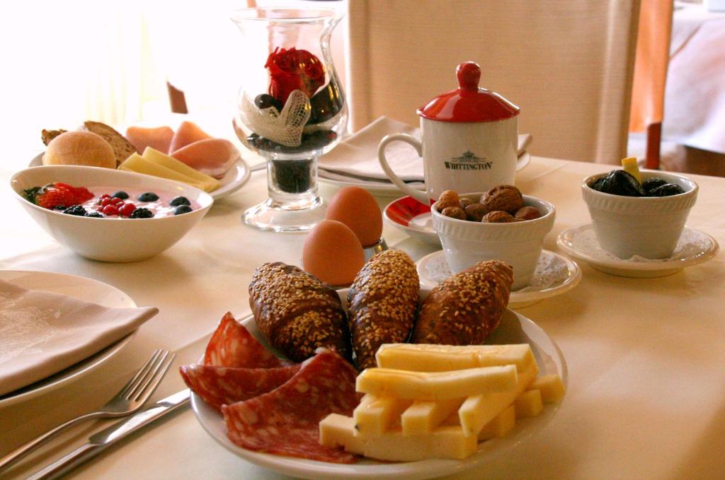 Bagātīga brokastu galda kompozīcija ar maizēm, sieru, desām, olām un augļu bļodiņu, kas ietver ogas. Uz galda atrodas arī rieksti un dažādi piedevas, radot sātīgu un veselīgu ēdienu izvēli.