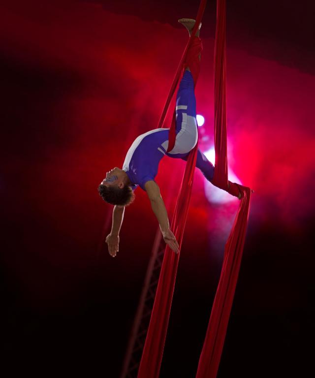 Gaisa akrobāts veic iespaidīgu triku, karājoties no sarkanām auduma lentēm, uz fona ar spilgtām gaismām.