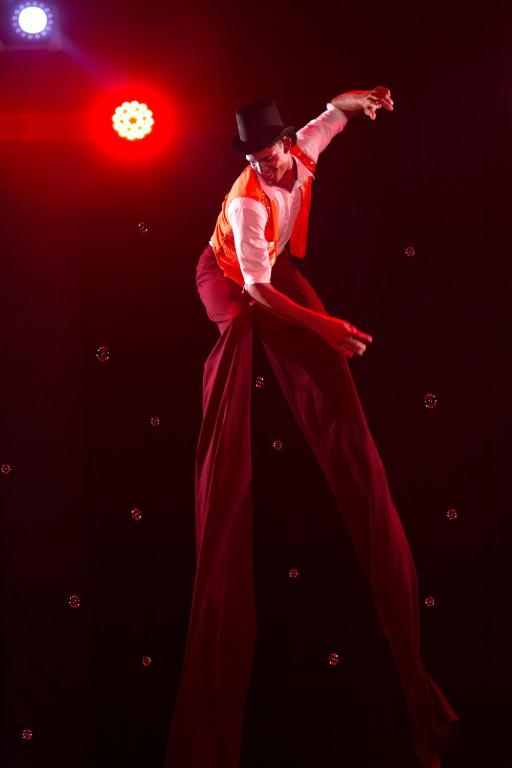 Augstāk stāvošs mākslinieks, kas ģērbies krāsainā kostīmā un cilindriskajā cepurē, izpilda akrobātiskus trikus uz gariem stiltos. Fons ir melns ar krāsainām gaismām, kas piešķir atmosfēru.