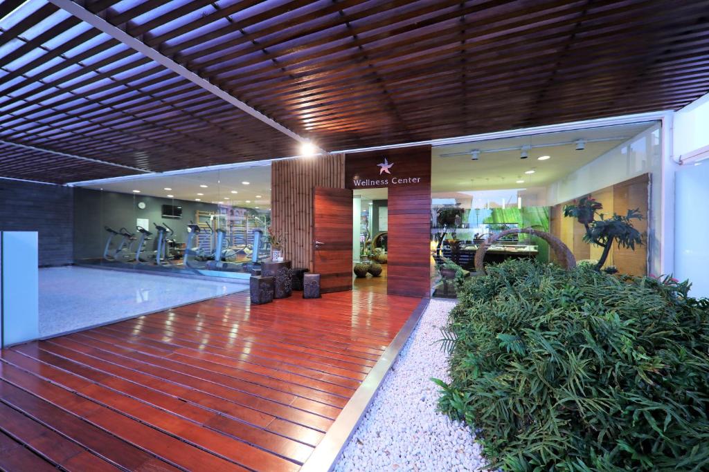 Mūsdienīgs wellness centrs ar elegantiem koka elementiem un zaļiem augiem, kas rada relaksējošu atmosfēru. Iekšpusē redzami sporta trenažieri un moderni interjera dizaina elementi.