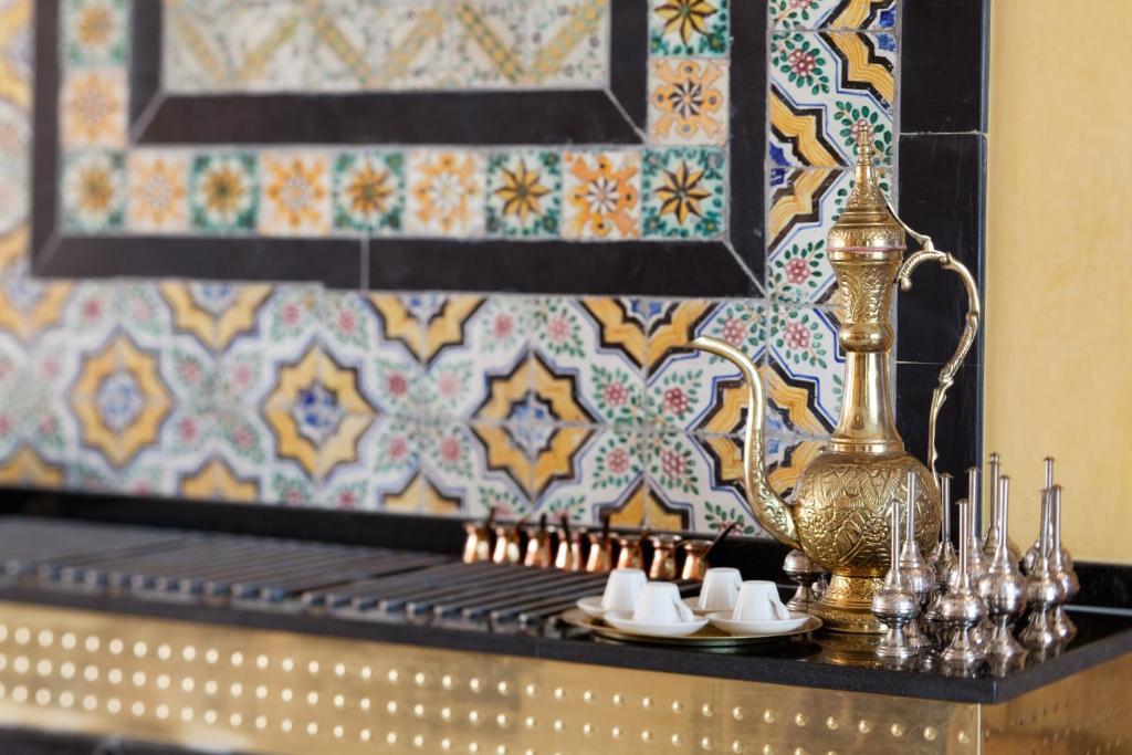 Misiņa tējkanna un elegants kafijas komplekts uz melna galda, ko rotā krāsains flīžu fons ar tradicionāliem rakstiem.
