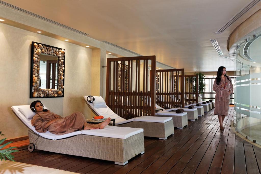 Spa telpa ar relaksācijas gultām, kurā viena sieviete klausās mūziku, bet otra staigā apkārt. Interjers ir elegants ar apgaismojumu un dekoratīviem elementiem.