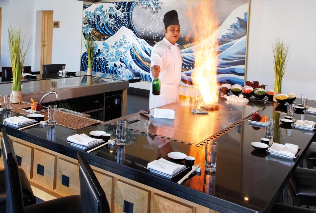 Šefpavārs veic ēdiena pagatavošanas triku ar flīsu liesmu, kamēr ap galdu ir redzami dažādi ēdieni un dekorācijas. Fons rotā gleznains vilnis, radot dinamisku atmosfēru.