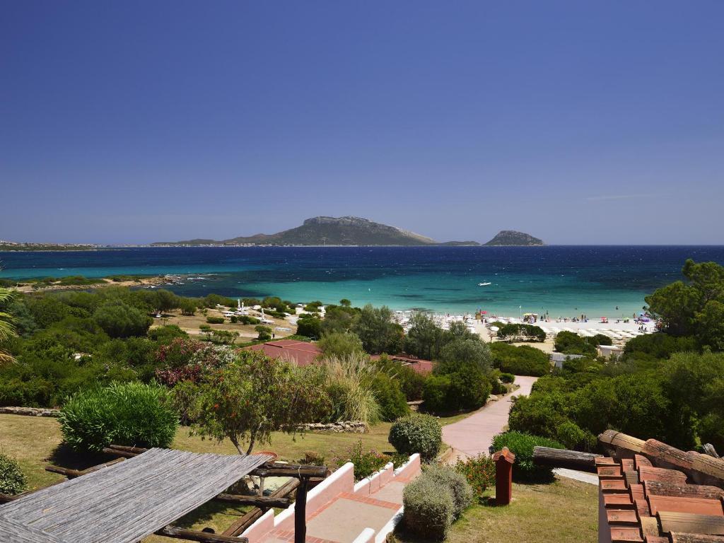 Skaists pludmales skats ar kristāldzidru ūdeni un zaļiem krūmiem, kas ieskauj pludmali. Tālumā redzami kalni un salas, kas piešķir ainavai pievilcību.