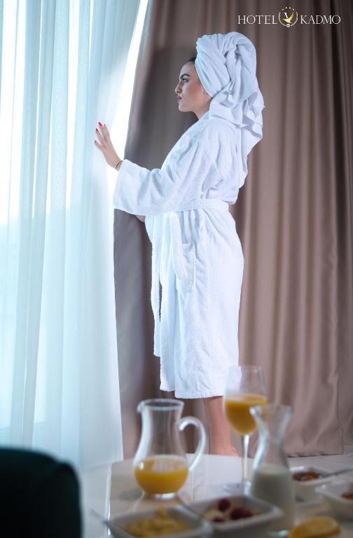 Sieviete baltā halātā, ar dvieli galvā, skatās pa logu, kamēr uz galda blakus ir brokastu ēdieni un dzērieni.