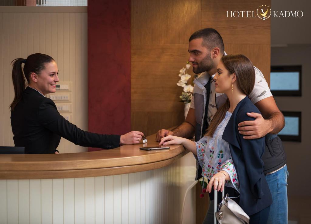 Reģistrācijas process viesnīcā, kur laipns darbinieks apkalpo pāri pie reģistratūras. Viesnīcas zīme "Hotel Kadmo" redzama augšējā stūrī.
