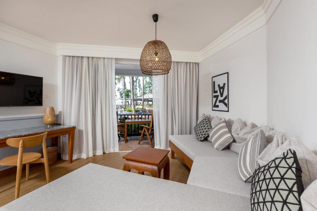 Mūsdienīga viesnīcas istaba ar pelēkiem dīvāniem, koka galdu un gaismas elementu, kas nodrošina mājīgu atmosfēru. Logi ved uz āru, kur redzami zaļumi un atpūtas zonas.