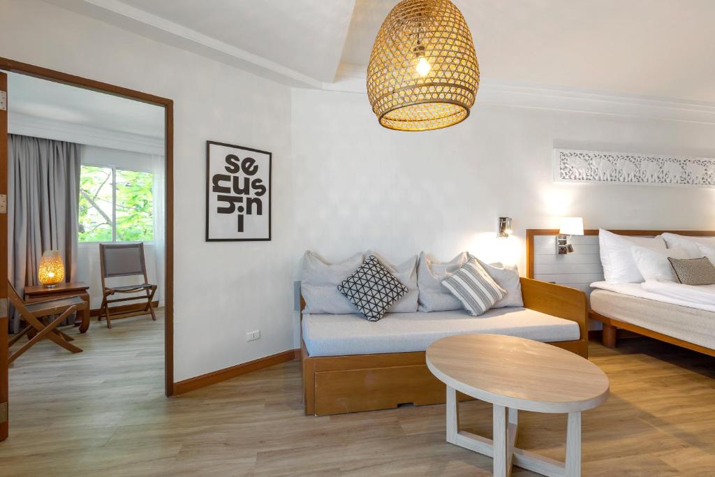 Mūsdienīgs viesnīcas numurs ar koka mēbelēm, gaišām krāsām un dabīgu apgaismojumu, kurā redzams dīvāns, gulta un dekoratīvas lampas. Numura interjerā ir mūsdienīga māksla un liels logs, kas piedāvā skatu uz ārpusi.