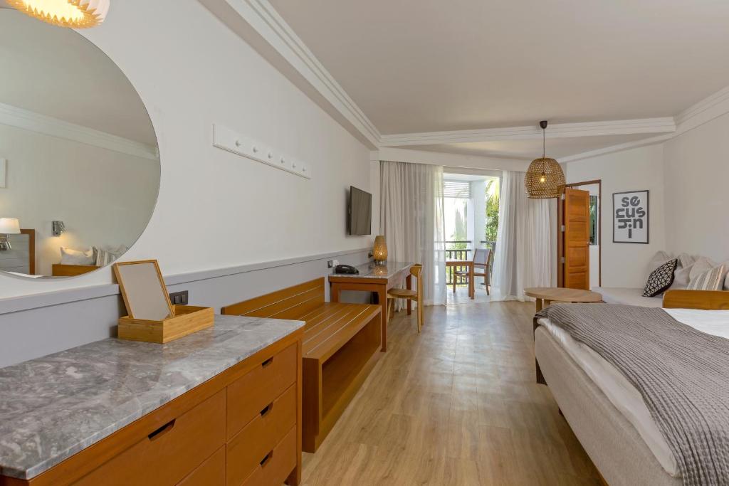 Mūsdienīga viesnīcas istaba ar gaišām krāsām, apaļu spoguli, koka mēbelēm un piekļuvi balkonam. Telpa ir eleganta un gaiša, radot relaksējošu atmosfēru.