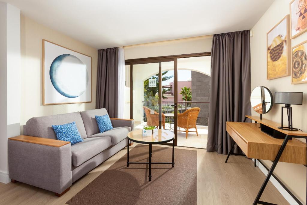 Mūsdienīgs viesistabas interjers ar pelēku dīvānu, dekoratīvām spilveniem un stikla galdu. Labi apgaismota telpa ar plašiem logiem, kas ved uz balkonu ar āra mēbelēm.