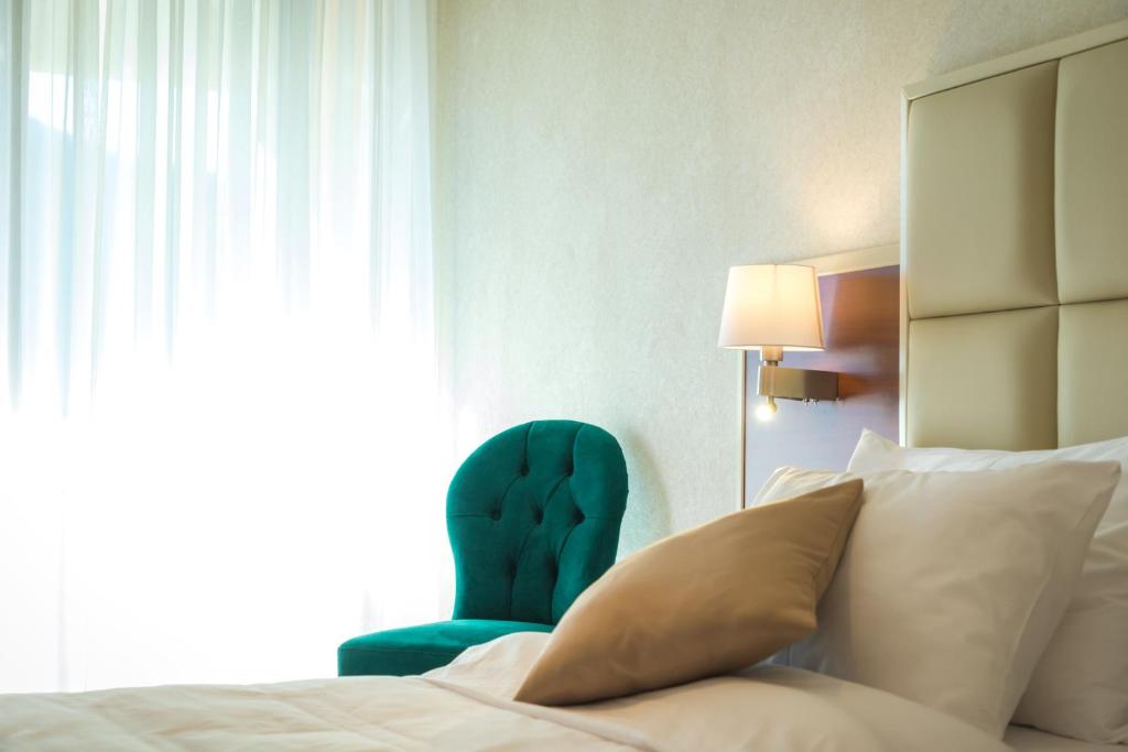 Mūsdienīgs guļamistabas interjers ar spilgtiem aksesuāriem, kas ietver zaļu krēslu un gaismas avotu. Gaisma plūst cauri caurspīdīgām aizkariem, radot mierīgu atmosfēru.