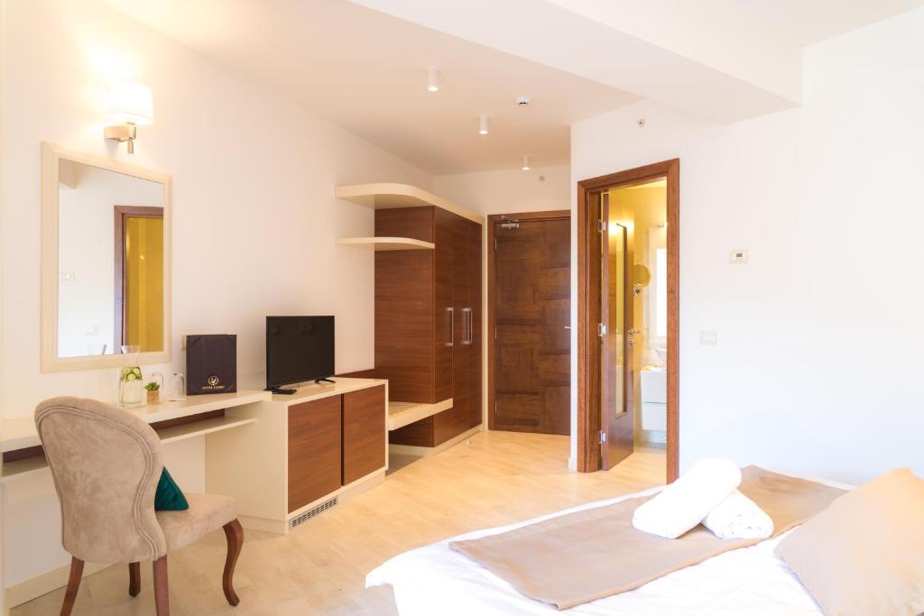 Moderns guļamistabas interjers ar gultu, rakstāmgaldu un televizoru, kas atrodas gaišā, eleganti iekārtotā telpā. Sienas ir izgaismotas ar iebūvētām lampām, un ir pieejami arī uzglabāšanas risinājumi.