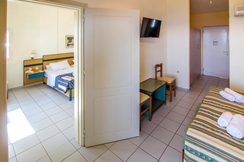 Numura iekārtojums ar gultu, galdu un krēslu, redzams caur atvērtām durvīm. Gaismas plūst iekšā no loga, radot viesmīlīgu atmosfēru.