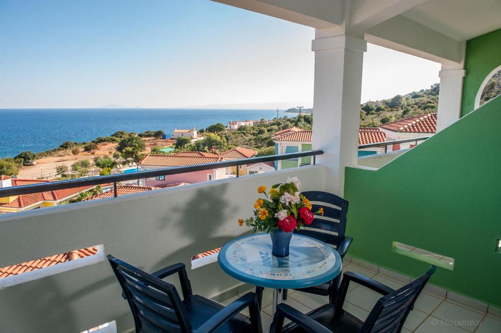 Skats no balkona ar galdu un krēsliem, uz kuru ir ziedu pušķis, pār skaistu jūras ainavu un krāsainām mājām.