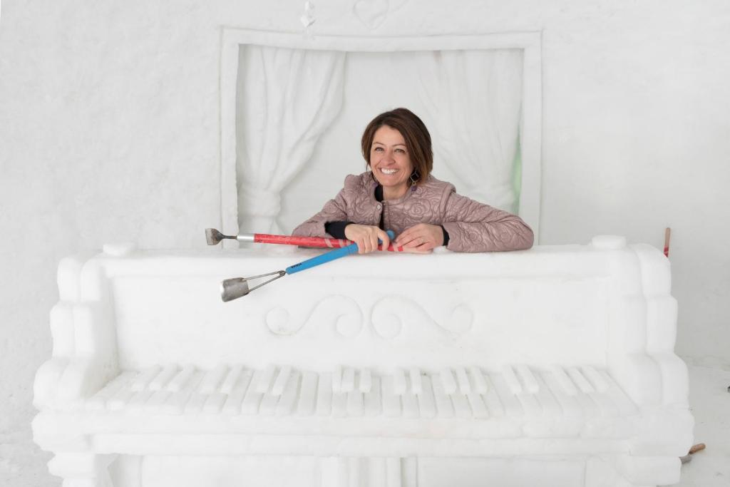 Sniega klavieru skulptūra ar sievieti, kas smaidot pozē, turot instrumentus. Sniega vide rada ziemas noskaņu un radošu atmosfēru.