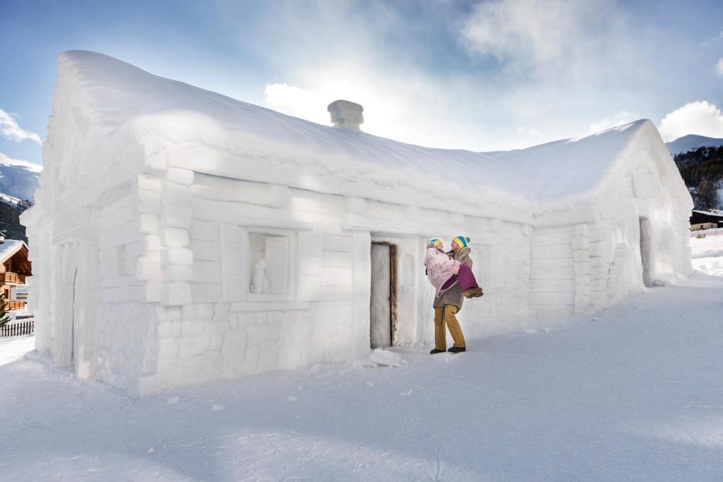 Sniega māja, kas izveidota no sniega, ar cilvēku, kurš tur bērnu. Apsnigušais ainava un zils debesis rada ziemīgu atmosfēru.