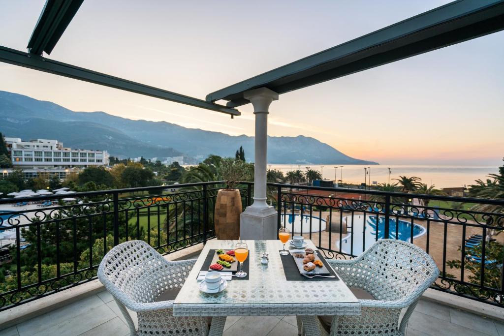 Brokastis uz balkona ar skatu uz jūru un kalniem, uz galda ir augļi, dzērieni un konditorejas izstrādājumi. Rīts ir mierīgs un saulains.