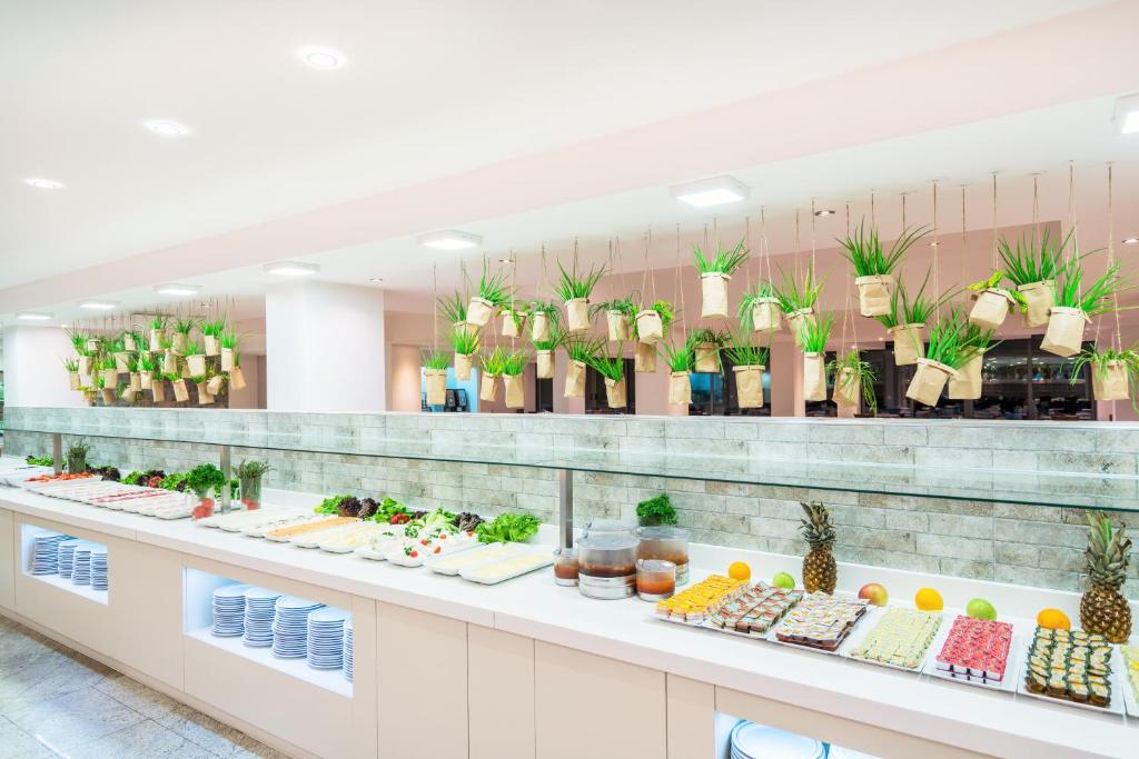 Bagātīgs bufetes galds ar dažādām salātiem, dārzeņiem un desertiem, kas papildināts ar stilīgiem, karājošiem augiem. Gaismas un modernais dizains rada patīkamu atmosfēru.