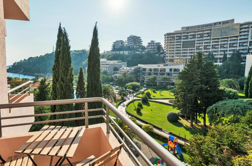 Skats no balkona uz zaļo teritoriju ar augstiem kokiem, viesnīcām un jūru fona. Gaisma apgaismo ainavu, radot mierīgu atmosfēru.