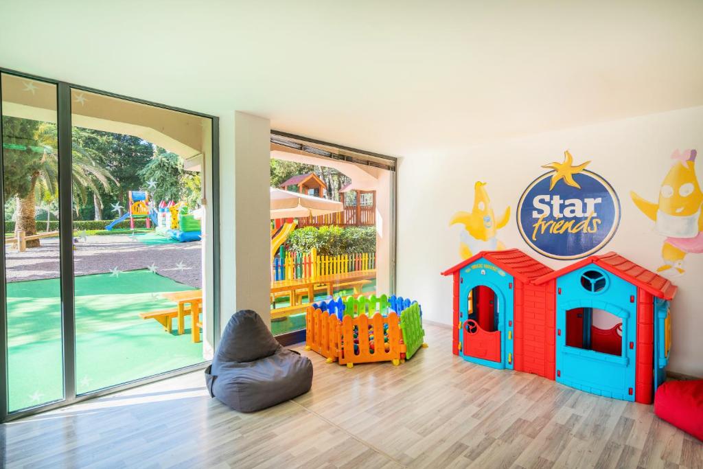 Krāsains bērnu rotaļu stūris ar plastmasas namiņiem, rotaļlietu žogu un mīksto krēslu. Logi paver skatu uz ārējo rotaļu laukumu ar zaļu zālienu un bērnu atrakcijām.