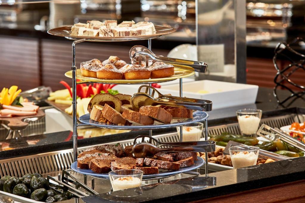 Daudzslāņu deserta statīvs ar dažādiem konditorejas izstrādājumiem, tostarp kūkām un maizītēm, izvietots pie bufetes. Fons satur augļus un citas uzkodas, radot bagātīgu brokastu atmosfēru.