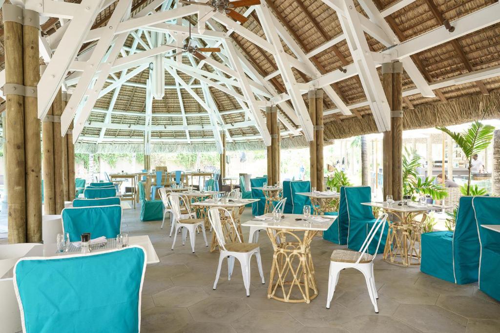 Elegants restorāns ar atvērtu telpu, kurā ir balti koka elementi un zilu sēdekļu pārvalki, radot vieglu un modernu atmosfēru. Apgaismojums un augi piešķir telpai dzīvīgumu.