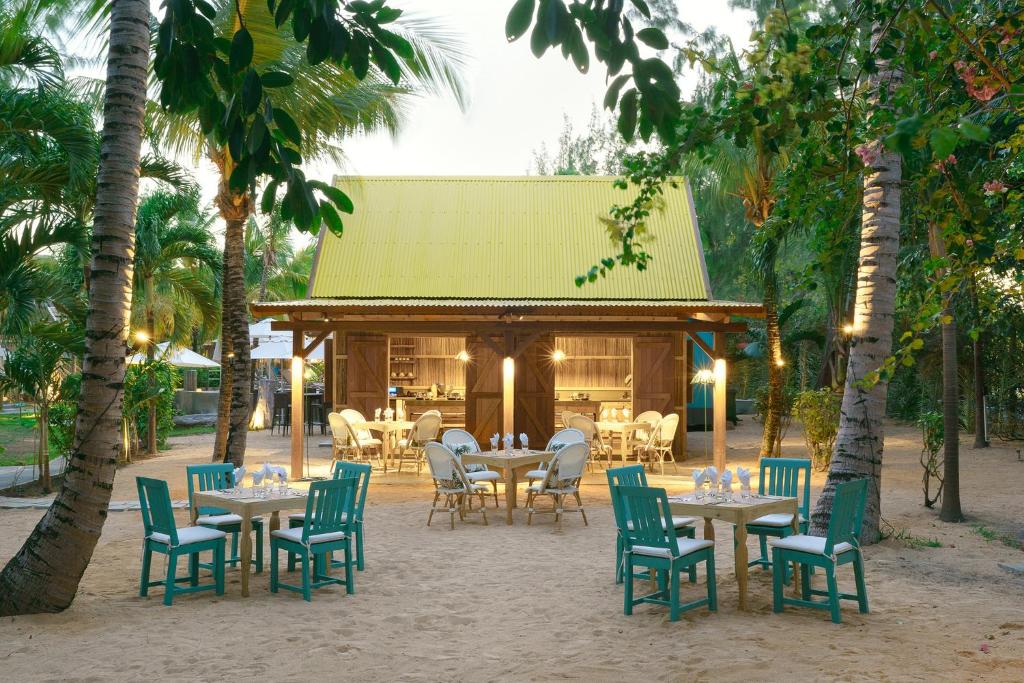 Tropu restorāns ar dzeltenu jumtu, apkārt palmām un smilšainu zemi, kurā atrodas zaļas krēsli un galdi. Ideāla vieta relaksācijai un ēdiena baudīšanai.