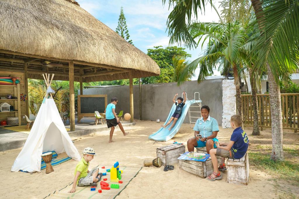 Bērnu spēļu laukums ar smilšu virsmu, kurā ir dažādas aktivitātes, piemēram, slidkalniņš, bumbu spēle un rotaļu laukums. Bērni un pieaugušie izklaidējas zem jumta, ko ieskauj zaļumi un palmas.