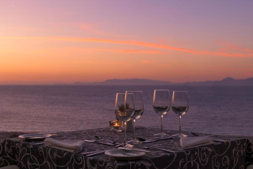 Romantisks skats uz saulrietu, kur uz galda ir noliktas glāzes un galda piederumi, radot ideālu atmosfēru vakariņām pie jūras.