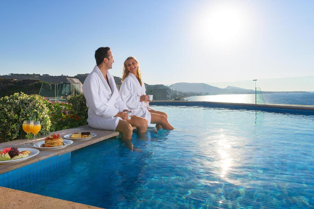 Pāris atpūšas pie baseina, ģērbti baltos halātos, baudot dzērienus un brokastis ar skatu uz jūru. Saules gaisma apgaismo ainavu, radot relaksējošu atmosfēru.