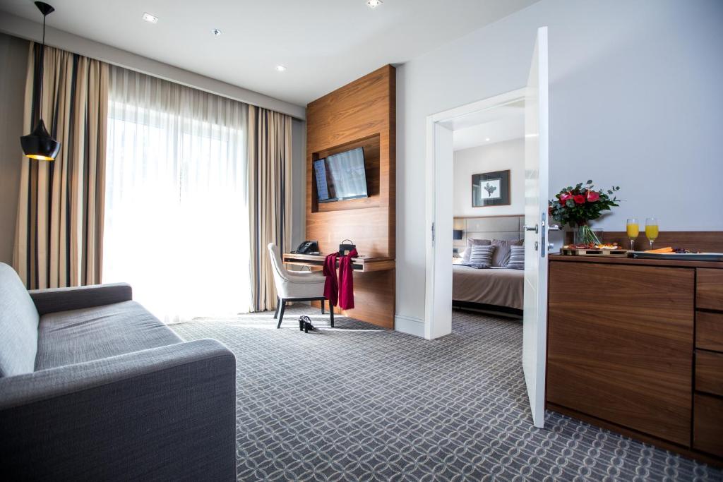 Mūsdienīga viesnīcas numura iekārtojums ar gaišām krāsām, dīvānu un darba zonu. Logu aizsedz eleganti aizkari, bet telpas stūrī redzama gulta un ziedu kompozīcija uz galda.