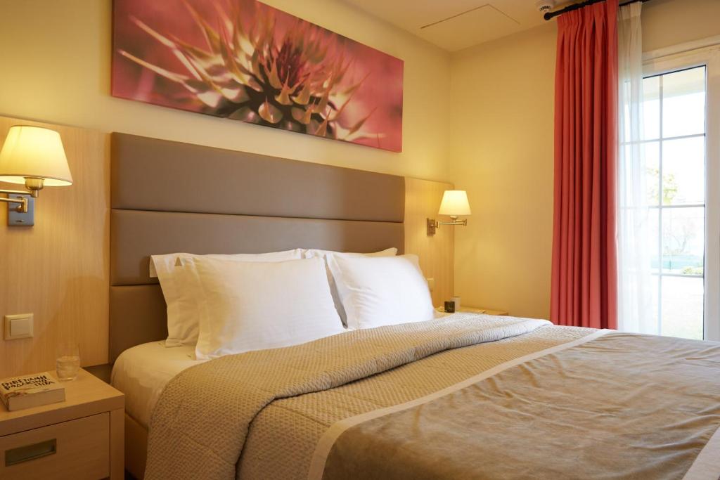 Mūsdienīgs guļamistabas interjers ar ērtu gultu, siltām krāsām un mākslas darbu uz sienas, kas piešķir telpai dzīvīgumu. Guļamistabā ir arī naktslampas un logi ar aizkariem.