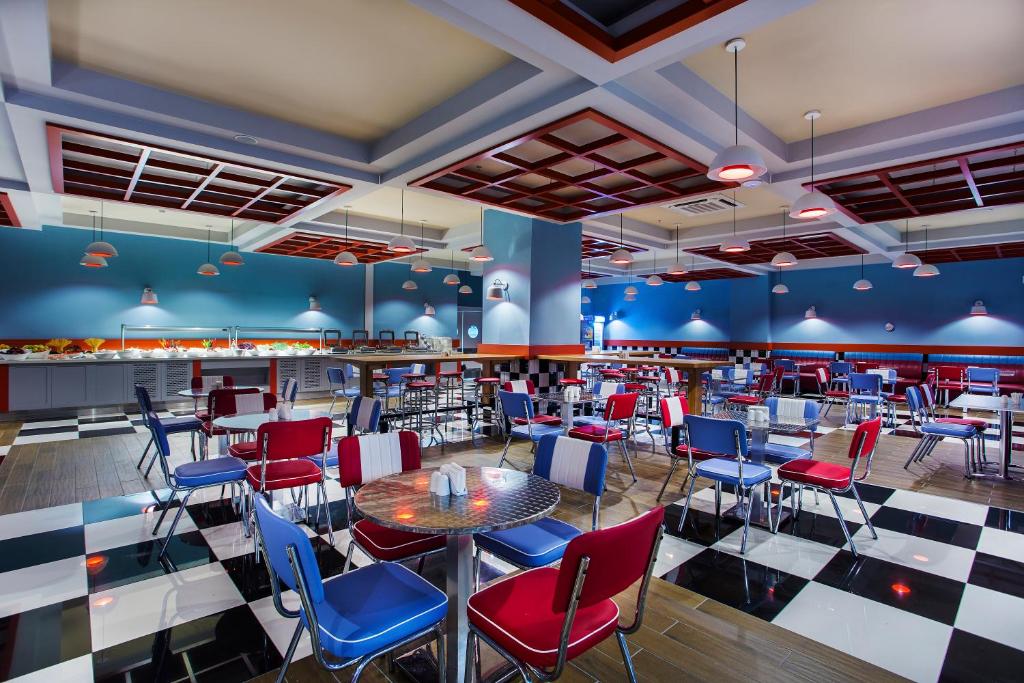 Mūsdienīga restorāna iekšskats ar krāsainām sarkanām un zilām krēslām, melnbaltu grīdu un stilīgu apgaismojumu. Telpa ir plaša un gaiša, radot patīkamu atmosfēru.