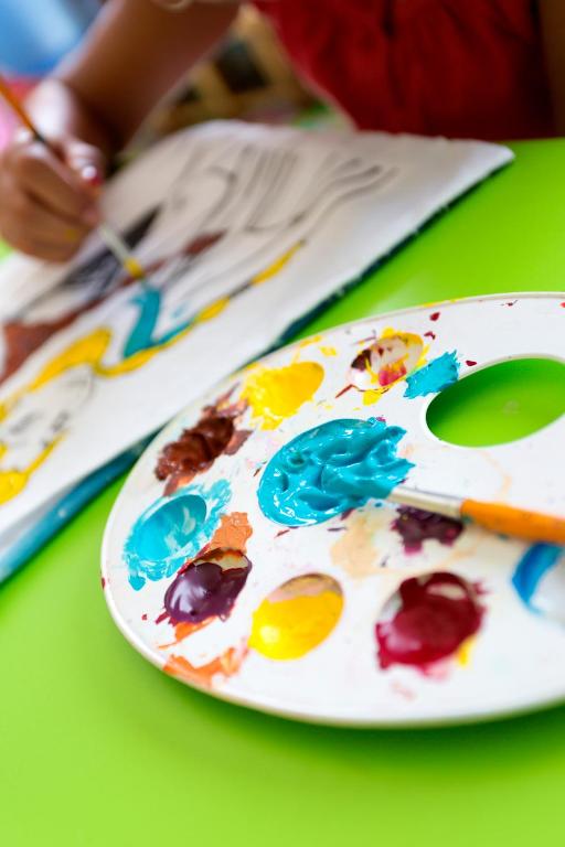 Bērns glezno, izmantojot krāsas uz paletes, kamēr fons ir zaļš. Krāsainas krāsas un gleznošanas process veido radošu atmosfēru.