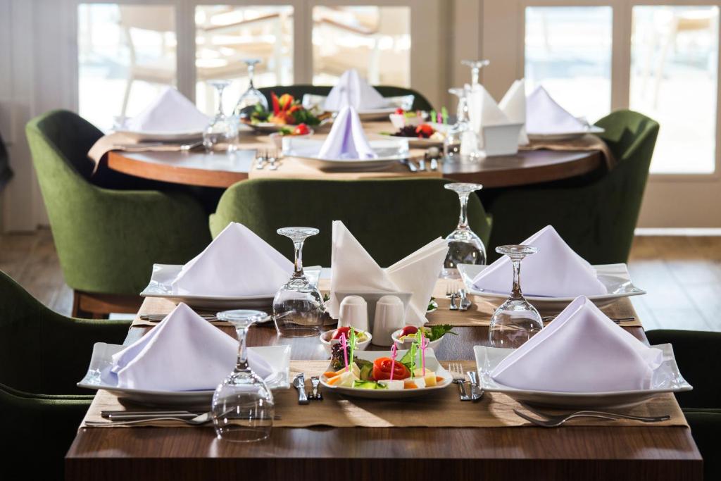 Eleganti izkārtots restorāna galds ar baltiem salvetēm, elegantiem traukiem un svaigiem salātiem, radot viesmīlīgu atmosfēru. Galds ir apņemts ar zaļām krēsliem un lieliem logiem, kas nodrošina dabiski apgaismojumu.