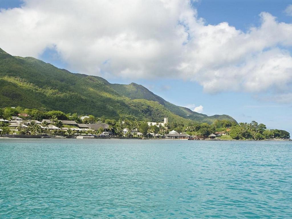 Skaists piejūras skats ar kalniem un zaļām palmām, kas veido idillisku tropu ainavu. Ūdens ir dzidrs un zils, radot relaksējošu atmosfēru.