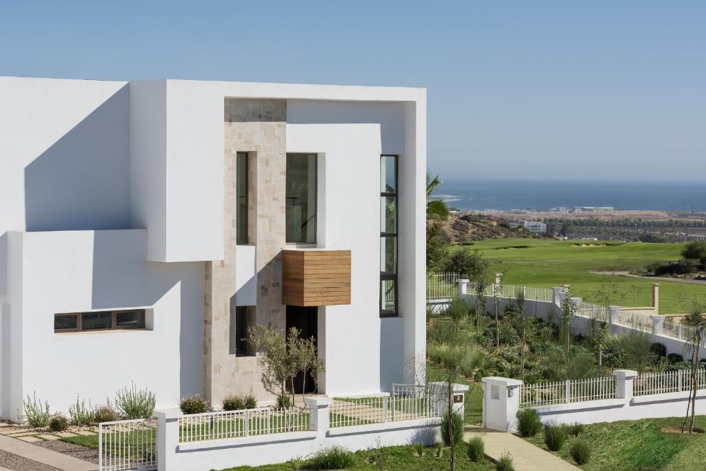 Mūsdienīga māja ar baltām sienām, koka detaļām un plašiem logiem, kas atrodas gleznainā ainavā ar skatu uz jūru un zaļo laukumu.