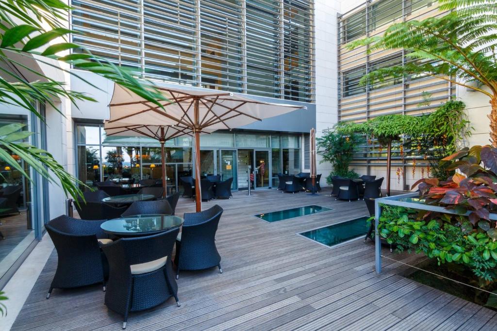 Mūsdienīgs terases interjers ar ērtām melnām krēsliem un stikla galdiem, apļveida saulessargiem un zaļām augu kompozīcijām, kas rada relaksējošu atmosfēru.