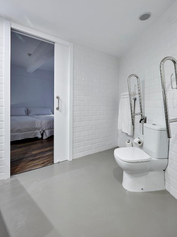 Mūsdienīga vannas istaba ar baltām flīzēm un minimālistisku dizainu, kas ietver tualeti un dvieļu turētāju. Aiz durvīm redzama guļamistaba ar gultu.