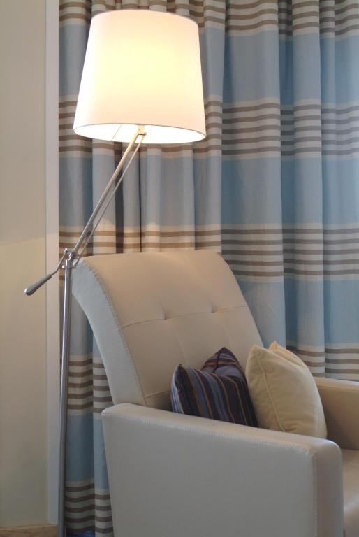 Mūsdienīgs krēsls ar spilveniem, blakus stāv lampa ar apaļu abatmentu, fonā ir svītraini aizkari.