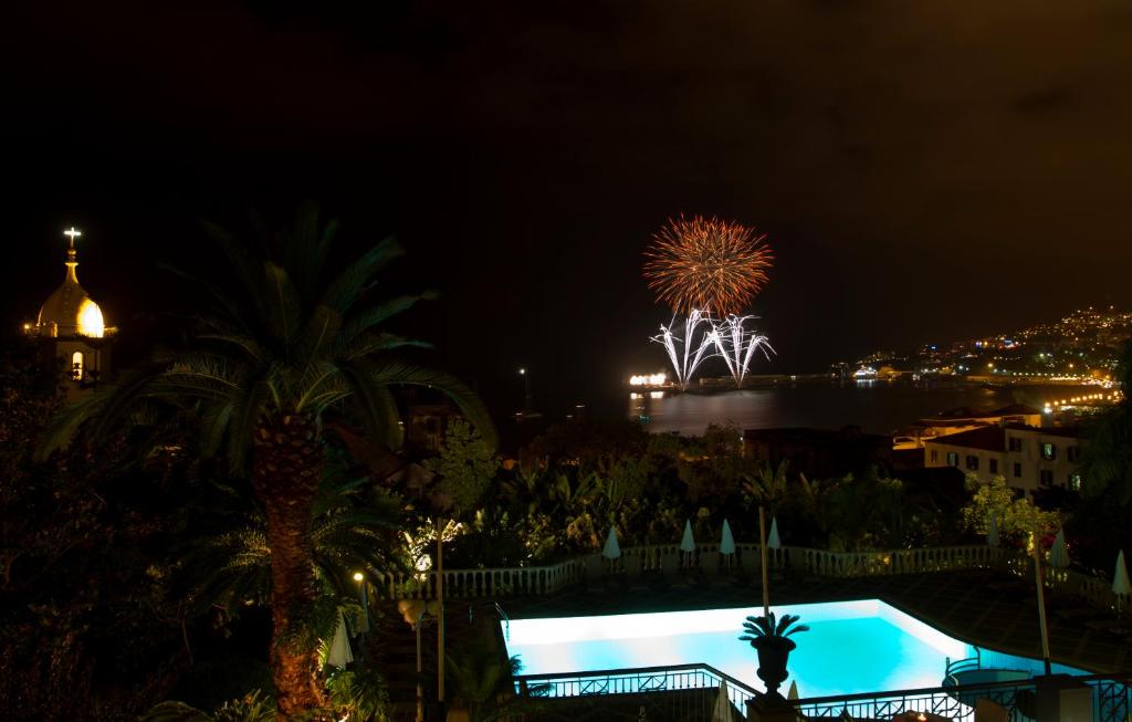 Naktī redzami krāšņi uguņošanas efekti, kas izgaismo debesis, ar jūras ainavu un baseinu priekšplānā. Fonā redzama pilsēta ar apgaismotām ēkām un palmām.