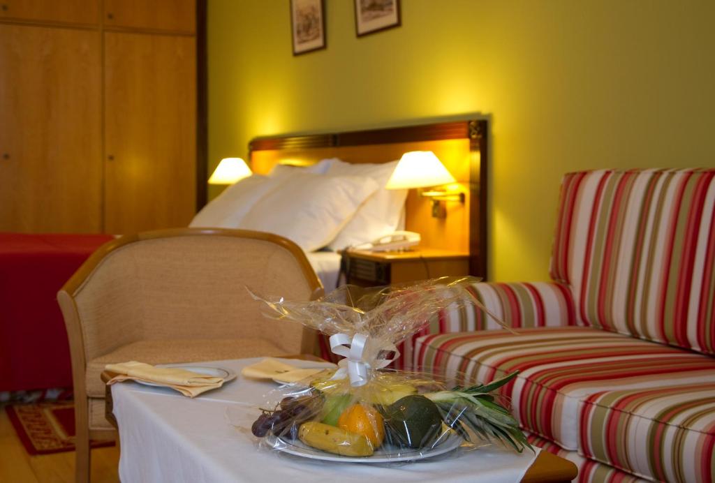 Mūsdienīga viesnīcas istaba ar spilgtu sienu, ērtiem krēsliem un augļu grozu uz galda. Redzama gulta un dekoratīvi elementi, kas rada patīkamu atmosfēru.