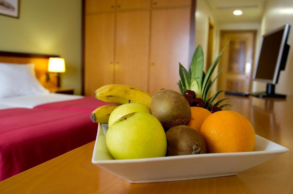 Augļu bļoda ar banāniem, āboliem, kivi, apelsīniem un vīnogām, novietota uz galda viesnīcas numurā ar gultu un televizoru fonā.