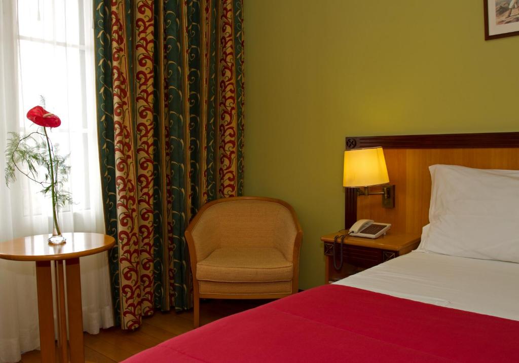 Mūsdienīga viesnīcas numura interjers ar sarkanu gultu, koka krēslu un dekoratīvām aizkaru drānām. Uz galda atrodas zieds vāzē, radot mājīgu atmosfēru.