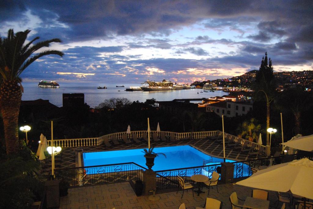 Nakts ainava ar baseinu un kuģiem ostā, apkārt palmām un apgaismojumu. Skats uz pilsētu un debesīm ar krāsainiem mākoņiem.