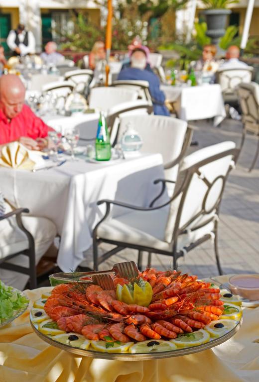 Lieliska garnelēm bagāta uzkodu plāksne ar citrona šķēlītēm un dārzeņiem, kas pasniegta elegantā restorāna terasē. Fons veido stilīgas galdiņas ar apkalpojošiem viesmīļiem un apmeklētājiem, kas baudīšanas brīdi.