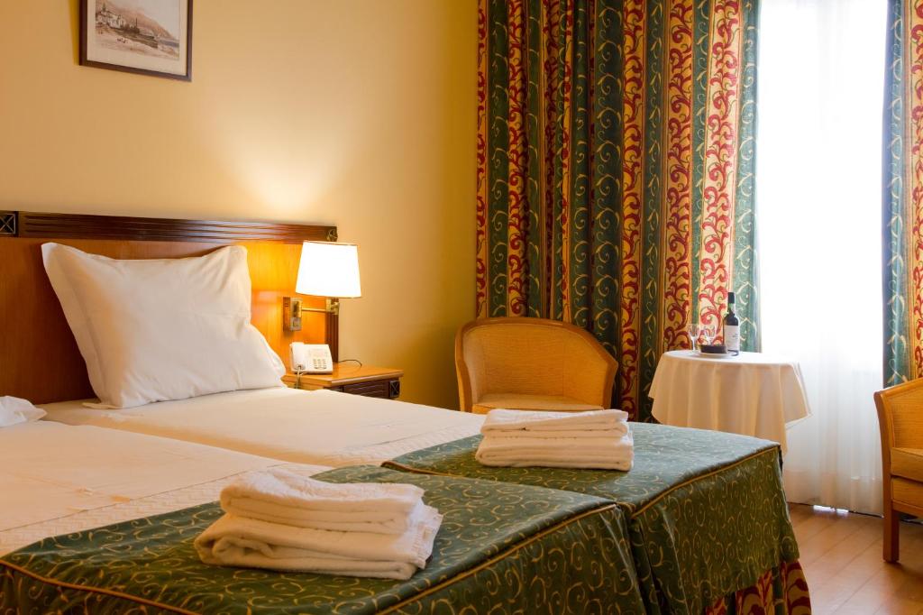 Mūsdienīgs viesnīcas numurs ar divām gultām, greznu aizkaru dizainu un siltu apgaismojumu. Uz galda atrodas vīna pudele un krēsls, radot patīkamu atmosfēru.