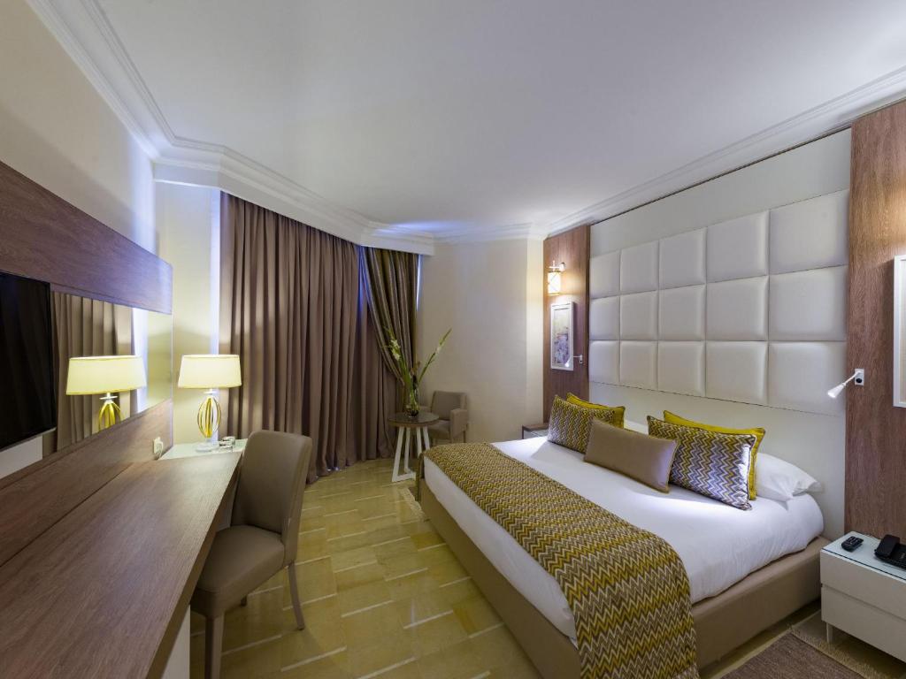 Mūsdienīgs viesnīcas numurs ar ērtu gultu un stilīgu interjeru, kas ietver koka un auduma elementus. Gaismas apgaismojums un logu aizkari rada patīkamu atmosfēru.