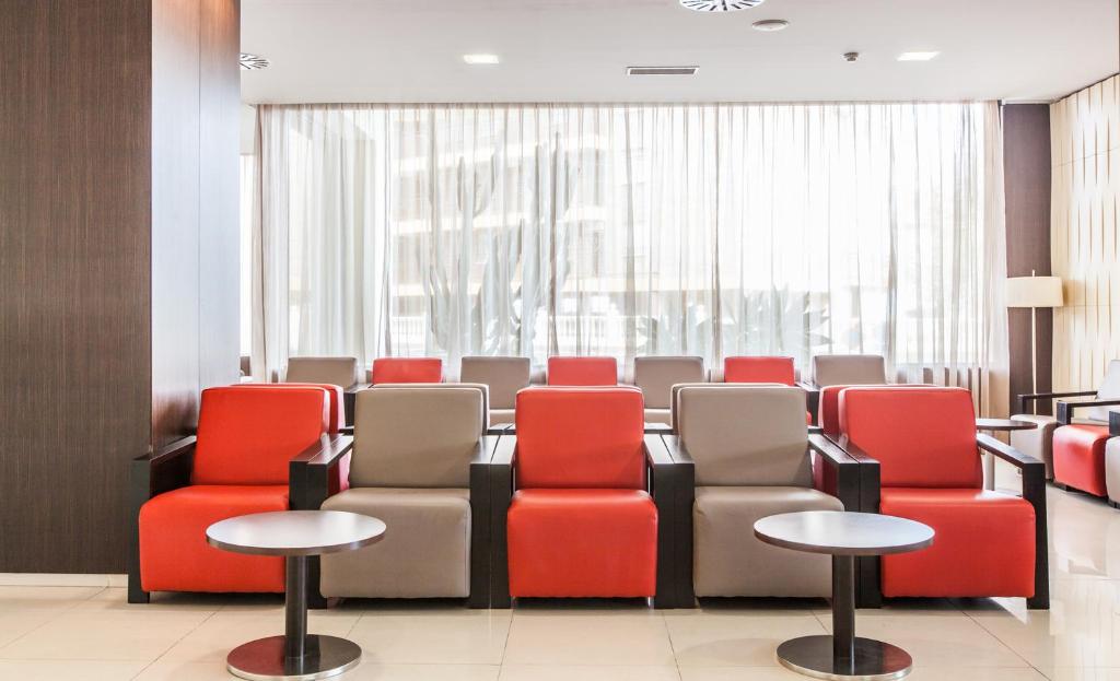 Mūsdienīga pieņemšanas zona ar sarkaniem un pelēkiem krēsliem un maziem galdiņiem, caur kuriem ieplūst dabiska gaisma.