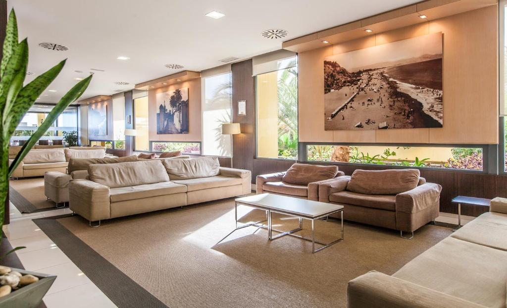 Mūsdienīgs viesnīcas uzgaidāmā telpa ar ērtām dīvānām, koka galdiem un mākslas darbiem uz sienām. Telpa ir gaiša un aicinoša, ar augiem un logiem, kas nodrošina dabisko apgaismojumu.